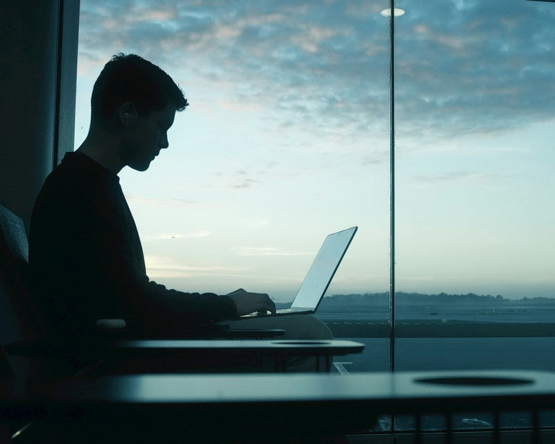 Person working on a laptop in an airport terminal with large windows showing a view of the sky.